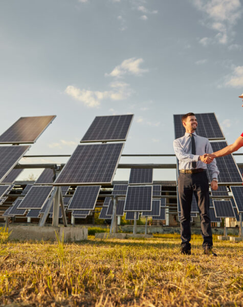 Supervisor and engineer shaking hands in solar power field Full body of male entrepreneur in formal wear making agreement with technician in uniform and hardhat in field with solar panels