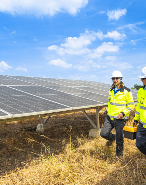 Two engineers wearing safety helmets and reflective jackets insp Two engineers wearing safety helmets and reflective jackets inspect a solar panel installation. One uses a drill tool, while the other checks the panel surface under a bright, sunny sky.