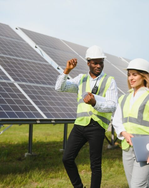Multiracial people having working discussion about green energy while standing outdoors. Solar panels arranged around. Multiracial people having working discussion about green energy while standing outdoors. Inspectors and foreman holding tablet, clipboard and drawings in hands. Solar panels arranged around.